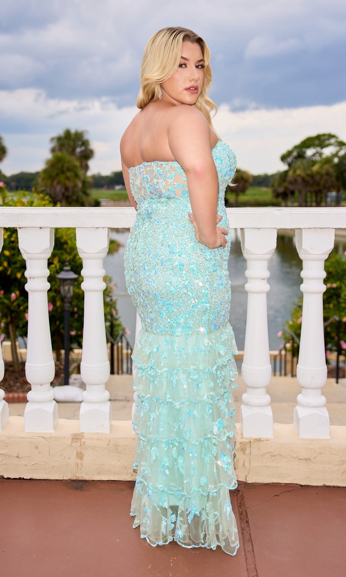 A woman in the Long Plus-Size Prom Dress: Primavera 14105, featuring a strapless neckline, light blue sequins, and a tiered ruffle skirt, stands on a terrace overlooking water with her back to the camera.